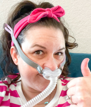 Mom giving a thumbs up while wearing CPAP machine wondering Why Is My Sleep So Bad Since Having Kids?