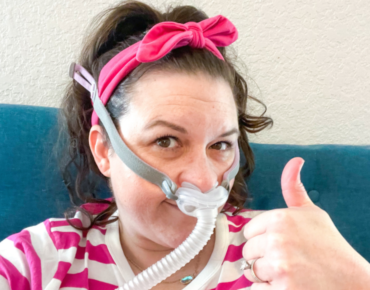 Mom giving a thumbs up while wearing CPAP machine wondering Why Is My Sleep So Bad Since Having Kids?
